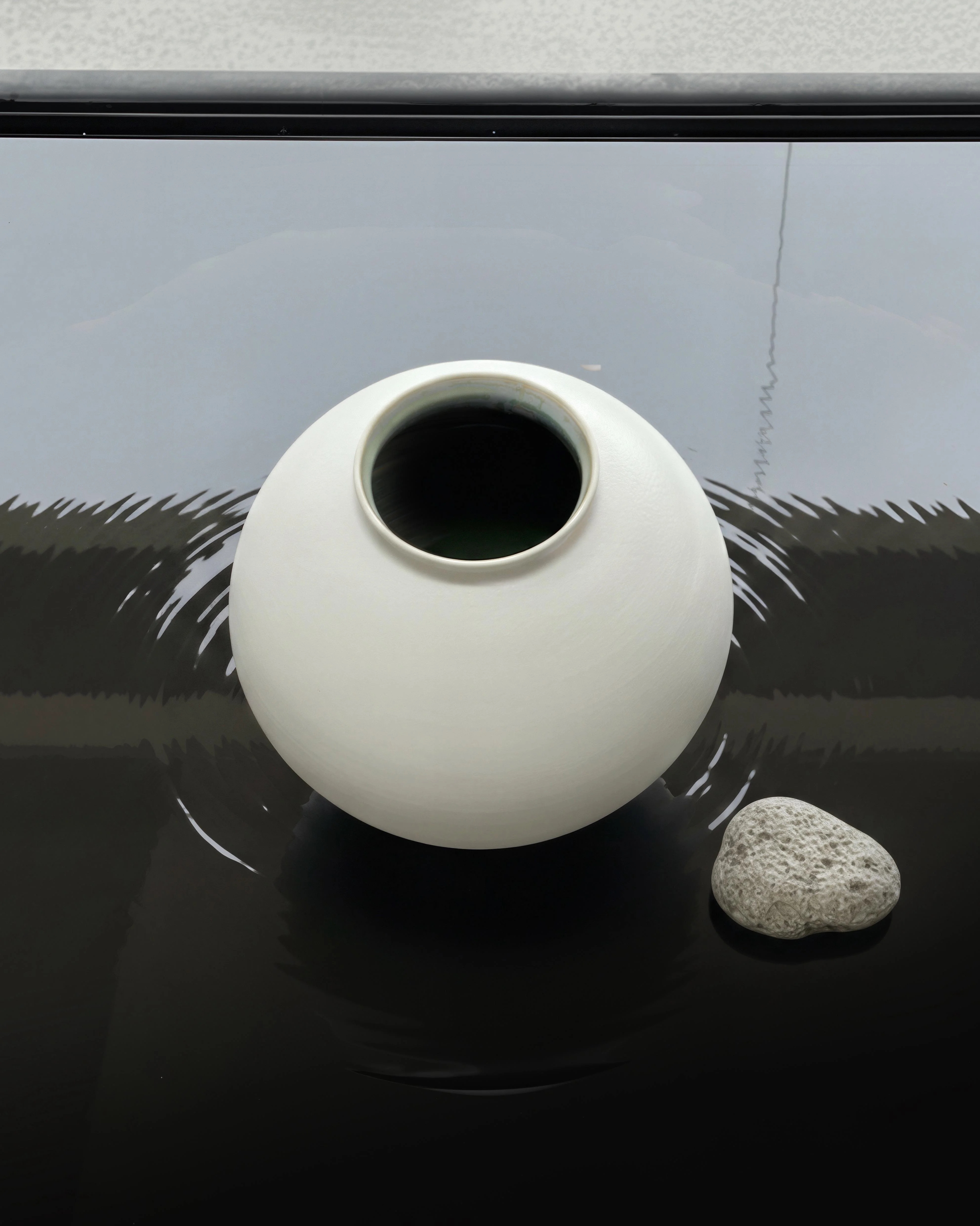 A white ceramic moon jar resting on a dark reflective surface beside a single stone